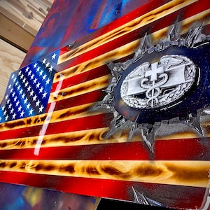 May include: A wooden American flag with a distressed, burnt look. The flag is painted in red, white, and blue. A silver medical insignia is embedded in the center of the flag.