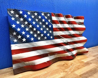 May include: A wooden American flag with a waving design. The flag features the traditional red and white stripes and a blue field with white stars. The wood grain is visible, adding texture to the patriotic artwork.