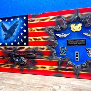 May include: A wooden American flag with a distressed look, featuring a black helicopter, a bald eagle, and military patches. The flag is painted with red, white, and blue colors and has a blue background with a text plaque.