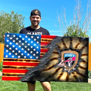 May include: A wood-burned American flag with a distressed look. The flag is split in half, with the top half showing the blue field of stars and the bottom half showing the red and white stripes. The right side of the flag is burned and cracked, revealing a black background with a stylized logo that says "FIST" in white letters. The logo is a shield with a fist holding a lightning bolt.