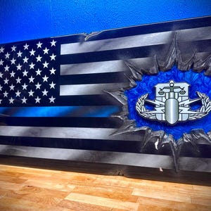 May include: A wooden American flag with a blue line, representing law enforcement, is broken through with a blue and white epoxy resin background. A silver military badge with a scale of justice, lightning bolts, and a wreath is centered in the resin.