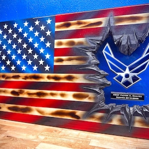 May include: Wooden American flag with a distressed design, featuring the US Air Force emblem. The flag has red and white stripes, a blue field with white stars, and a carved-out section. Includes text: SSGT Victor A. Chavez, Served 2016-2025.
