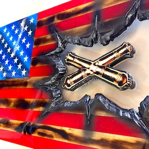 May include: A wooden American flag with a distressed look, featuring a raised, clear epoxy resin section with a gold crossed cannons emblem. The flag is painted in red, white, and blue.