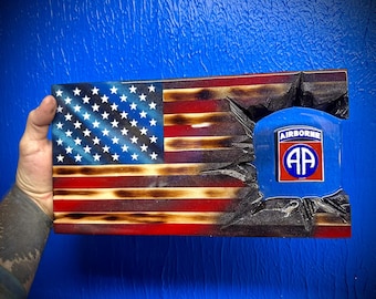 May include: A wooden American flag with a distressed, burnt look. The flag is partially torn, revealing a blue patch with the 82nd Airborne Division insignia, which reads "AIRBORNE" and features a red circle with the letters "AA".