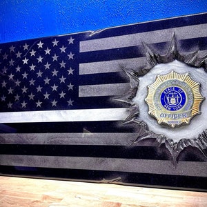May include: A black and white American flag with a thin blue line, a police badge, and the words "Corrections Community Supervision State of New York Officer 60849" are visible in the center of the flag.