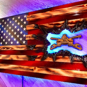 May include: A wooden American flag with a distressed look, featuring a blue and gold Army Corps of Engineers emblem. The flag is lit with blue LED lights.