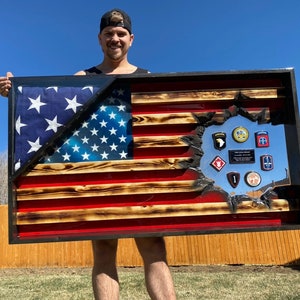 May include: A wooden American flag with a distressed look, featuring a cutout section with military patches and a silver background. The flag is framed in black and is being held by a person.
