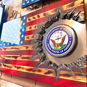 May include: Wooden American flag with red and white stripes and a blue field of stars. The center features the Department of the Navy seal, set within a distressed, textured border. The flag is a patriotic home decor item.