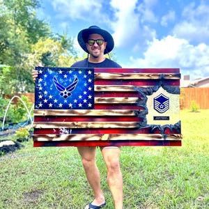 May include: A person holding a wooden American flag with a blue Air Force emblem and a burnt wood effect. The flag also features a military rank insignia with the text "Master Sergeant" and "USAF".