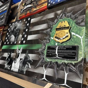 May include: A black and white American flag with a green stripe across the bottom. The flag is decorated with a gold badge that says "CBP Border Patrol U.S. Patrol Agent" and a plaque that reads "In Honor Carlos I. Pereyra".