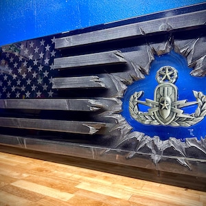 May include: Wooden American flag with a distressed design. The flag features a blue field with white stars and gray stripes. A blue circle reveals a military emblem. The flag is mounted on a wooden surface.