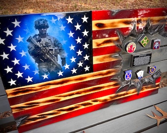 Pode incluir: Uma bandeira americana em estado de deterioração com um soldado em camuflagem segurando um rifle na frente do campo azul de estrelas. A bandeira está parcialmente rasgada e tem uma variedade de patches militares e uma placa com texto no lado direito.