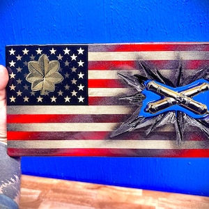 May include: A wooden American flag with a gold leaf design and crossed cannons in the center. The flag is painted in red, white, and blue with a distressed look.