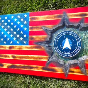 May include: A wooden American flag with a distressed look, featuring a blue and white star field and red and white stripes. The flag has a circular emblem in the center, with the text "United States Space Force Department of the Air Force MMXIX".