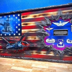 May include: A wooden American flag with a distressed look, featuring a black helicopter breaking through the flag. The flag is framed with a black wood frame. The right side of the flag is broken away to reveal a blue background with a silver military patch, a silver pin, and a silver badge. The badge reads "Night Stalkers".