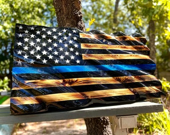 May include: A wooden American flag with a blue line running horizontally across the middle. The flag is made of wood that has been burned and then sealed with a clear epoxy resin. The flag is displayed on a wooden shelf.