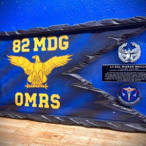 May include: A wooden plaque with a blue background and a gold eagle emblem. The text "82 MDG" and "OMRS" are in gold. Includes a silver emblem and a plaque with text: "LT COL KIERAN DHILLON".