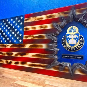 May include: Wooden American flag with a distressed design. The flag features red and white stripes, blue field with white stars, and a blue emblem with gold lettering and the text "PRO DEO ET PATRIA". The name "CH(CPT) JASON E. MCGILL" is also visible.