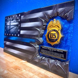 May include: A wooden plaque featuring a distressed American flag design with a blue line, a US enforcement badge, and text that reads "SPECIAL AGENT CASEY RETTIG". The plaque has a textured, dimensional appearance with a blue background.