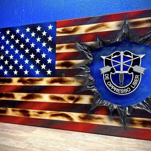 May include: A wooden American flag with a distressed, burnt look. The flag has a blue circle with a silver emblem in the center. The emblem has a dagger, arrows, and the text "DE OPPRESSO LIBER".