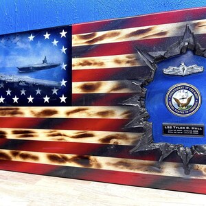 May include: A wooden American flag with a distressed, burnt look. The flag has a hole in the center, revealing a blue background with a Navy seal and a plaque that reads "LS2 Tyler C. Hull".