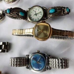 May include: Four wristwatches with different colored faces and metal bands. The watches have silver, gold, and blue bands. The faces are white, gold, and blue.