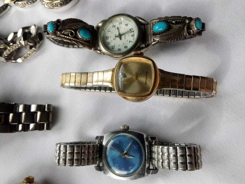 May include: Four wristwatches with different colored faces and metal bands. The watches have silver, gold, and blue bands. The faces are white, gold, and blue.