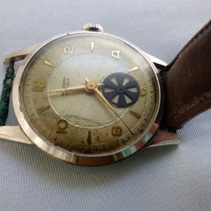 May include: A vintage wristwatch with a silver case and a brown leather band. The watch face has a white background with black Roman numeral hour markers and a black center wheel.