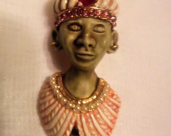 May include: A green and white figurine of a person wearing a turban with a red jewel on top. The figurine is wearing a white and pink collar with a gold and pearl necklace. The figurine is holding a white pearl.