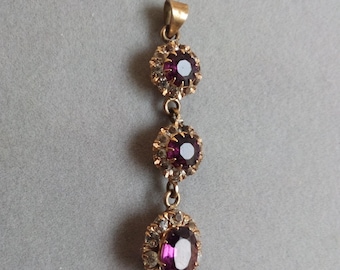 May include: A gold-toned pendant with three purple gemstones. The top and middle stones are round, while the bottom stone is oval. Each gemstone is surrounded by smaller, clear stones.