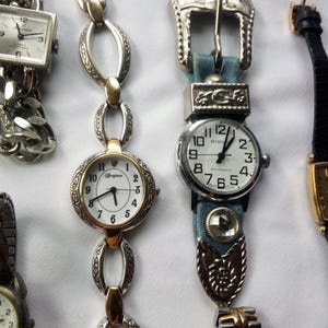 May include: Five wristwatches with different styles and designs. The watches have silver, gold, and black bands. One watch has a blue denim band with a silver buckle.