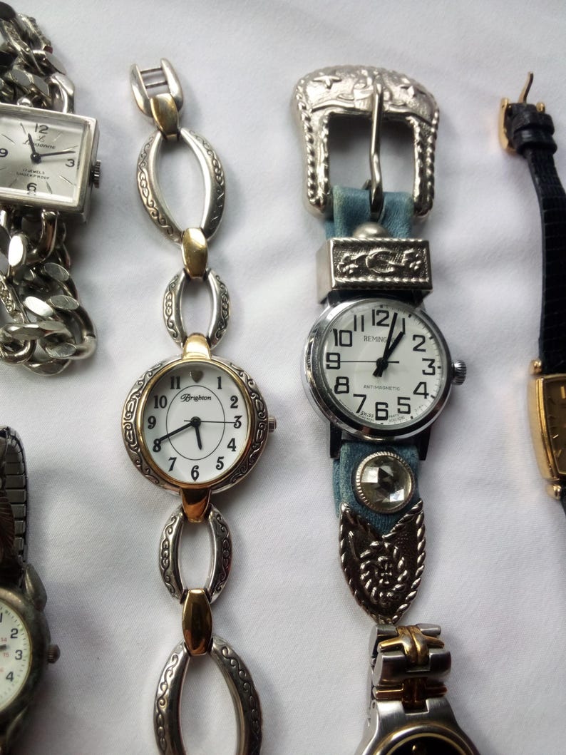 May include: Five wristwatches with different styles and designs. The watches have silver, gold, and black bands. One watch has a blue denim band with a silver buckle.