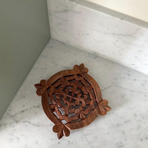 Puede incluir: Un salvamanteles redondo de madera tallada intrincadamente con un diseño floral. El salvamanteles está hecho de madera marrón oscuro y presenta un patrón calado detallado. Cuatro elementos decorativos en forma de hoja están colocados alrededor del borde.