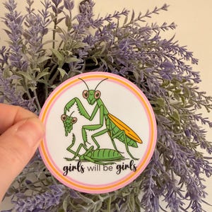 May include: A round sticker featuring a green praying mantis illustration with orange wings and the text "girls will be girls". The sticker has a pink and orange border and is held in front of a lavender wreath.