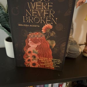 May include: A book titled You Were Never Broken by Melissa Acosta with a black cover featuring gold text and an illustration of a person with long red hair adorned with sunflowers. The subtitle reads A funny, supportive, and spiritual guide.