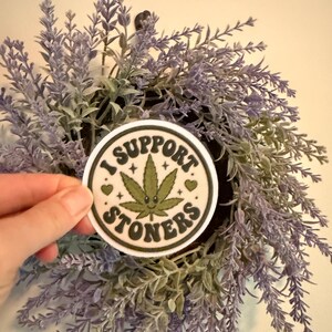 May include: A round sticker with the text "I SUPPORT STONERS" encircling a smiling green cannabis leaf. The sticker is held in front of a lavender and green floral wreath.