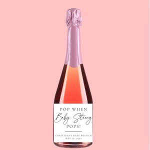 May include: A white label with black text that reads "Pop When Baby Strong Pops!" and "Christina's Baby Brunch May 15, 2023" is attached to a pink champagne bottle.