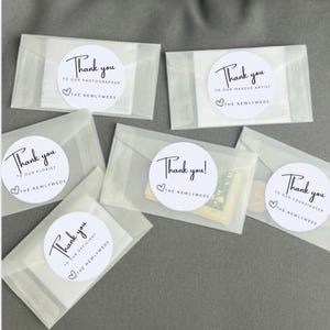 May include: Several small, translucent envelopes with white circular stickers. The stickers read "Thank you" followed by different titles like "photographer" and "coordinator", with a heart and "THE NEWLYWEDS" below. The envelopes are arranged on a gray surface.