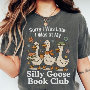 Pode incluir: T-shirt cinzenta escura com o texto "Sorry I Was Late I Was at My Silly Goose Book Club". A t-shirt apresenta um gr&aacute;fico de tr&ecirc;s gansos de desenho animado carregando livros e caf&eacute;, com detalhes florais.