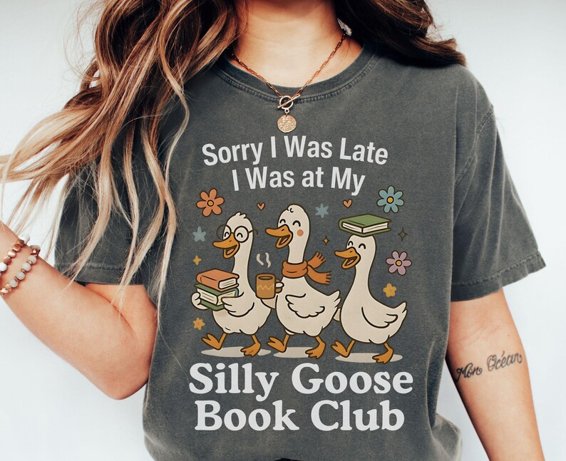 Pode incluir: T-shirt cinzenta escura com o texto "Sorry I Was Late I Was at My Silly Goose Book Club". A t-shirt apresenta um gr&aacute;fico de tr&ecirc;s gansos de desenho animado carregando livros e caf&eacute;, com detalhes florais.