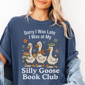 Pode incluir: Camiseta azul marinho com o texto "Sorry I Was Late I Was at My Silly Goose Book Club". O gr&aacute;fico apresenta tr&ecirc;s gansos de desenho animado carregando livros e caf&eacute;. Acentos florais e estrelas circundam os gansos.