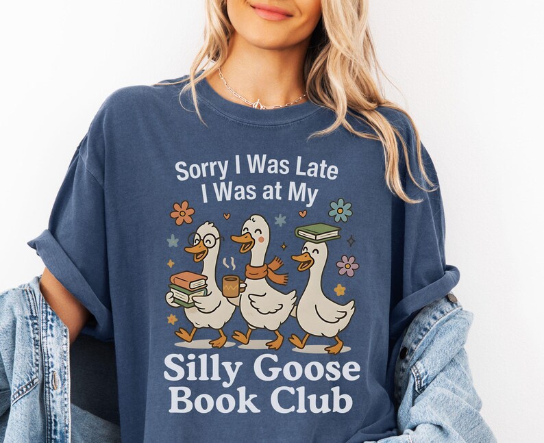 Pode incluir: Camiseta azul marinho com o texto "Sorry I Was Late I Was at My Silly Goose Book Club". O gr&aacute;fico apresenta tr&ecirc;s gansos de desenho animado carregando livros e caf&eacute;. Acentos florais e estrelas circundam os gansos.
