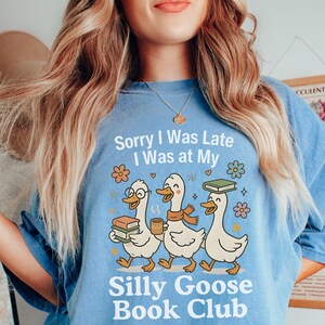 Pode incluir: Camiseta azul clara com o texto "Sorry I Was Late I Was at My Silly Goose Book Club." O desenho apresenta tr&ecirc;s gansos de desenho animado com livros e caf&eacute;. Os gansos usam &oacute;culos e cachec&oacute;is. A camiseta tem um corte folgado.