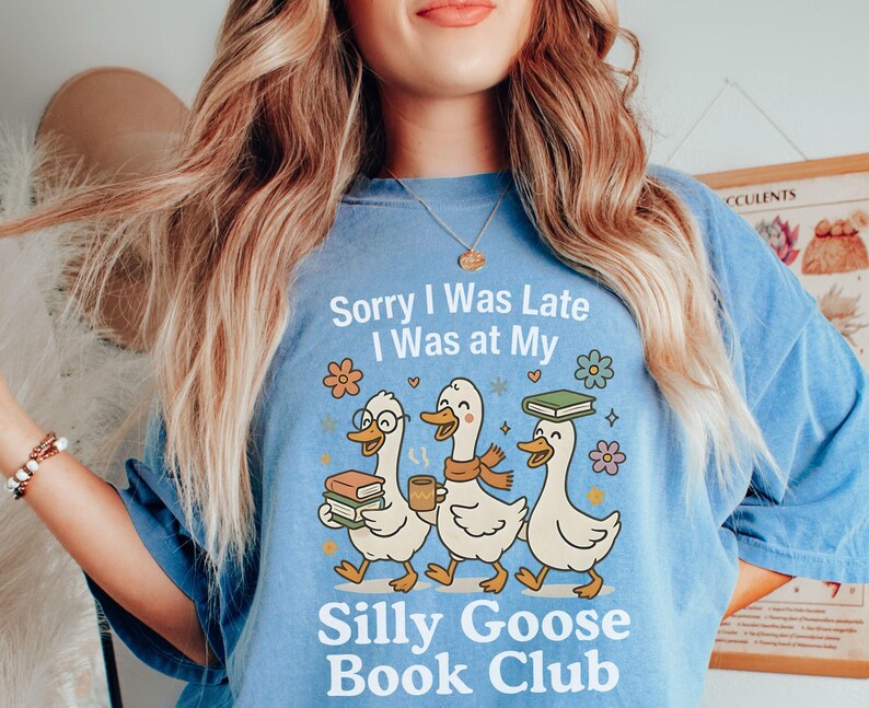Pode incluir: Camiseta azul clara com o texto "Sorry I Was Late I Was at My Silly Goose Book Club." O desenho apresenta tr&ecirc;s gansos de desenho animado com livros e caf&eacute;. Os gansos usam &oacute;culos e cachec&oacute;is. A camiseta tem um corte folgado.