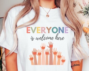 May include: White t-shirt featuring the phrase "EVERYONE is welcome here" in various colours. Below the text are illustrations of hands holding hearts. A casual, everyday wear item.