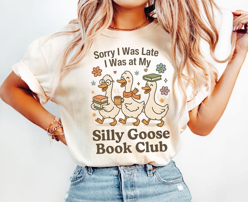 May include: Cream-colored t-shirt with the text "Sorry I Was Late I Was at My Silly Goose Book Club." The graphic features three cartoon geese carrying books and coffee, with floral accents. The shirt is made of a soft material.