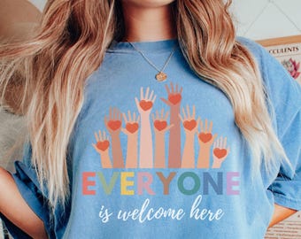 May include: A light blue t-shirt featuring a graphic of hands reaching up with hearts in their palms. The text "EVERYONE is welcome here" is printed below the graphic.