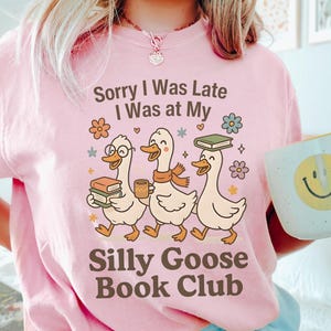 Pode incluir: T-shirt rosa claro com o texto "Sorry I Was Late I Was at My Silly Goose Book Club". O design apresenta tr&ecirc;s gansos de desenho animado com livros e caf&eacute;. Uma caneca com um smiley tamb&eacute;m &eacute; vis&iacute;vel.