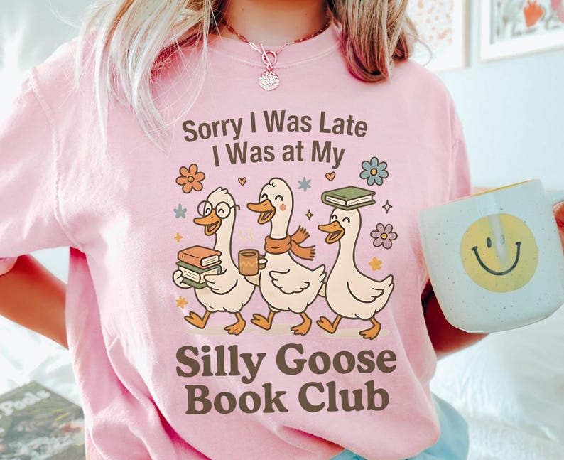 Pode incluir: T-shirt rosa claro com o texto "Sorry I Was Late I Was at My Silly Goose Book Club". O design apresenta tr&ecirc;s gansos de desenho animado com livros e caf&eacute;. Uma caneca com um smiley tamb&eacute;m &eacute; vis&iacute;vel.