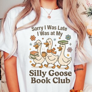 Pode incluir: Camiseta branca com o texto "Sorry I Was Late I Was at My Silly Goose Book Club". O design apresenta tr&ecirc;s gansos de desenho animado, cada um com livros ou uma x&iacute;cara de caf&eacute;, com motivos florais e estrelas.